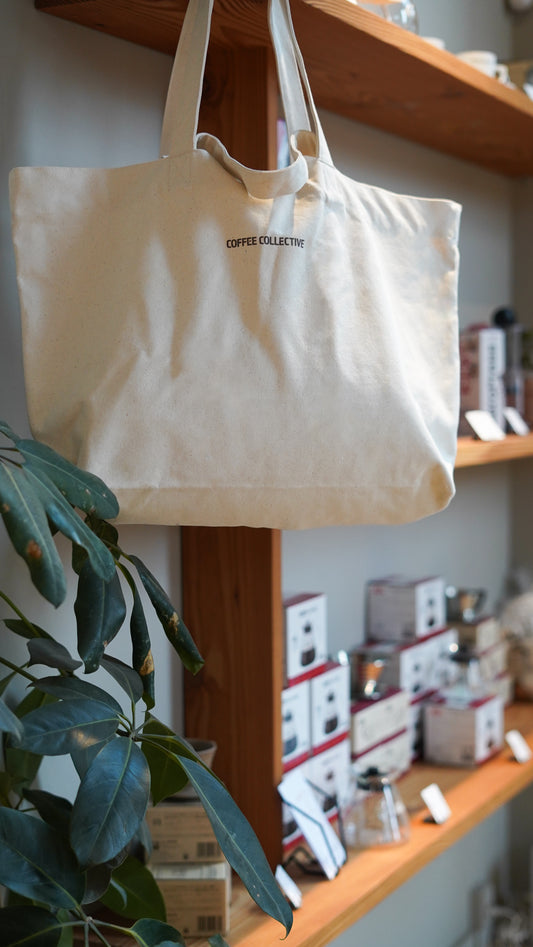 Beige tote bag with 'Coffee Collective' branding hanging on a wooden shelf.