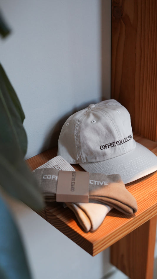 Beige cap with 'Coffee Collective' text and a pair of branded socks on a wooden shelf