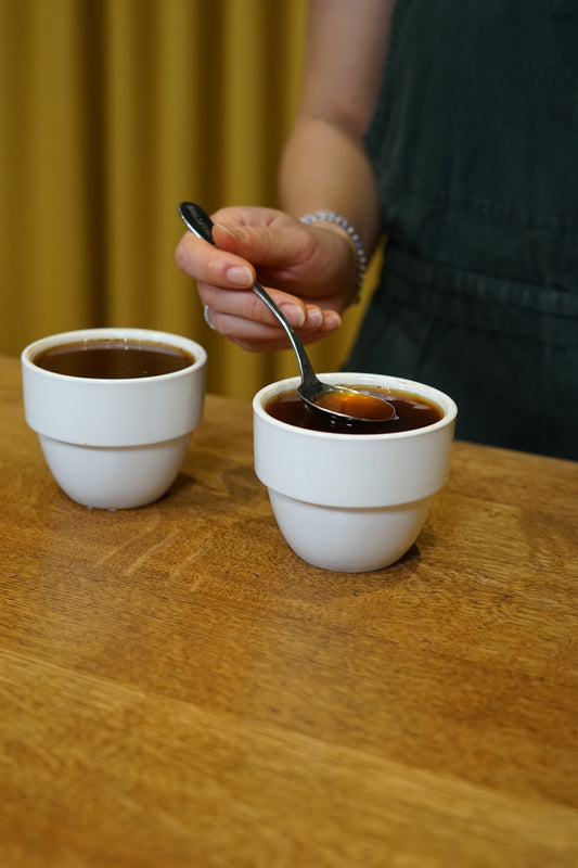 Two white cups on a wooden surface with a hand holding a spoon, scooping from one of the cups.